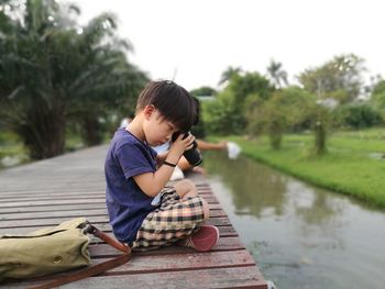 Side view of boy looking through binoculars sitting on boardwalk against sky