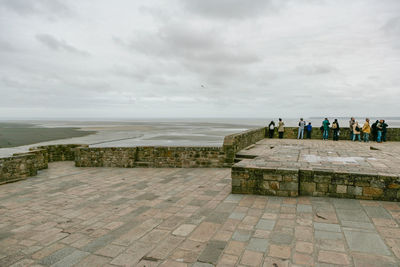 People on footpath by sea against cloudy sky