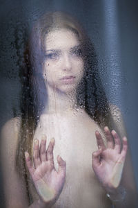 Portrait of shirtless woman looking through wet glass window