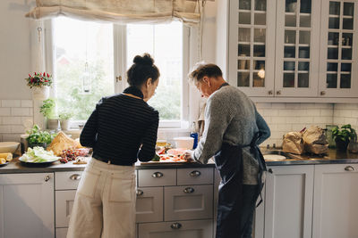 Active female and male senior friends cutting watermelon at counter in kitchen