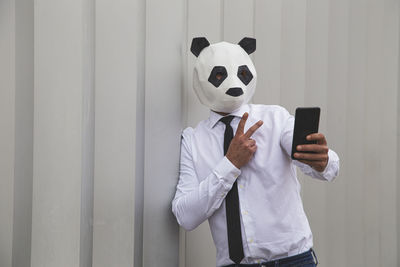 Man in white shirt, tie and panda mask taking smartphone selfie against wall