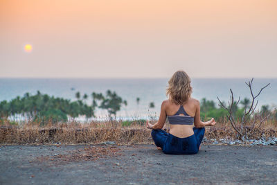 Rear view of woman practicing yoga on footpath against sea during sunset