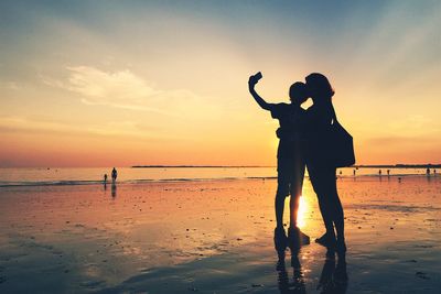 Silhouette friends taking selfie while standing at beach against sky during sunset