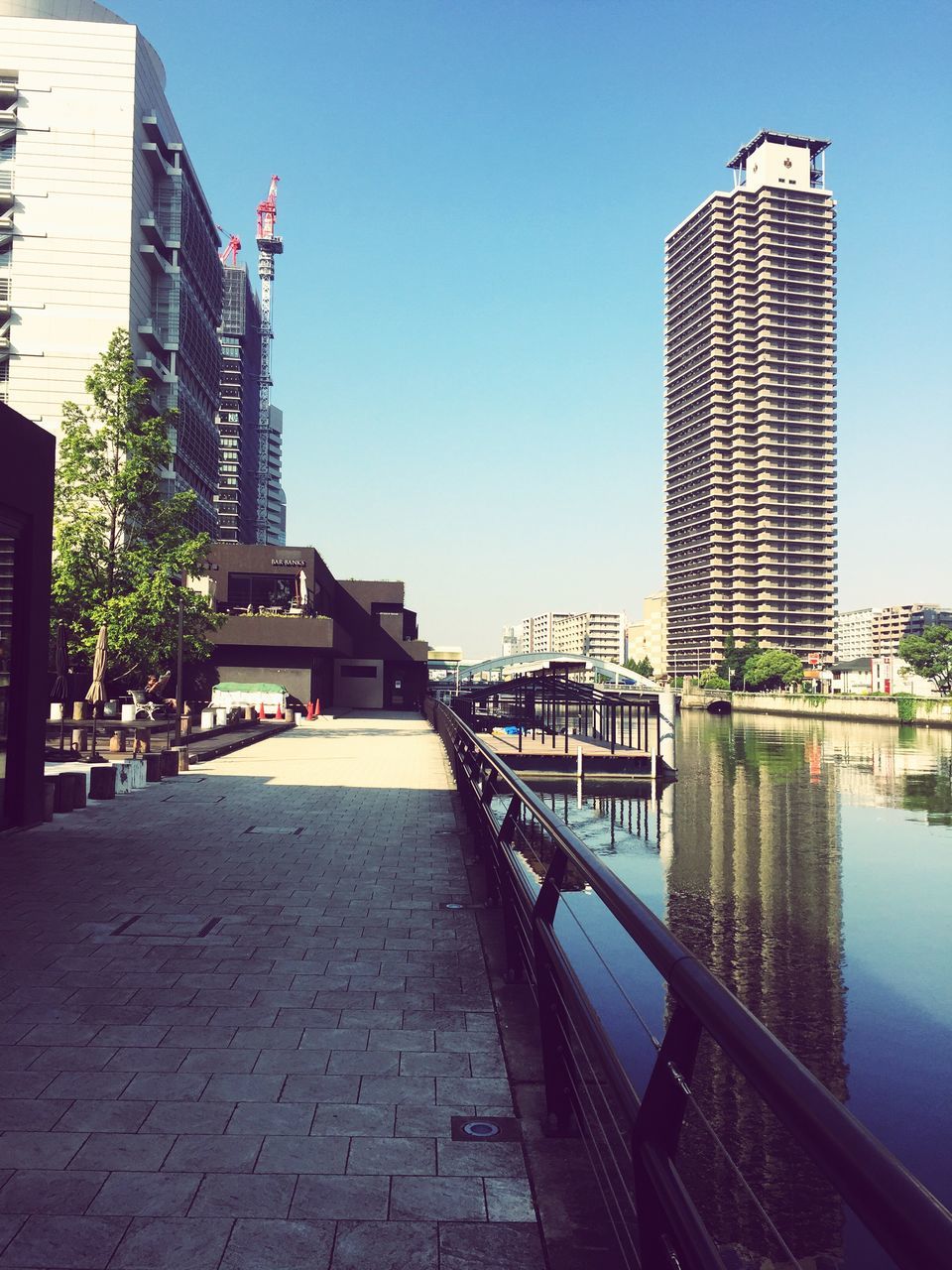 architecture, building exterior, built structure, clear sky, the way forward, city, water, sunlight, railing, blue, walkway, building, day, incidental people, outdoors, diminishing perspective, footpath, tower, sky, sidewalk