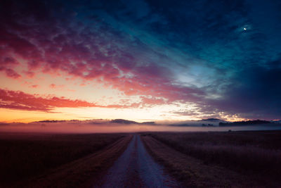 Empty dirt road on field against sky during sunset