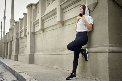 Full length of jogger looking away while standing on sidewalk against wall in city