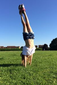 Full length of girl doing handstand on grassy field against clear blue sky