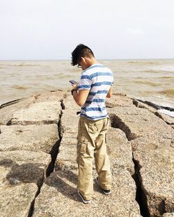 Rear view of man standing with mobile phone on rocks by sea against sky