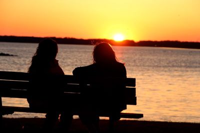 Rear view of women sitting on bench at beach during sunset