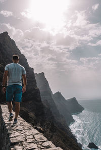 Rear view of man standing on cliff by sea against sky