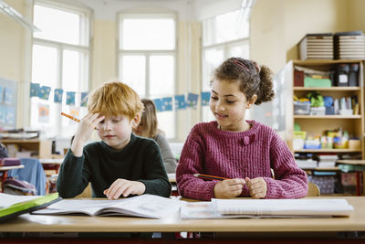 Smiling girl sitting by confused blond boy at desk in classroom