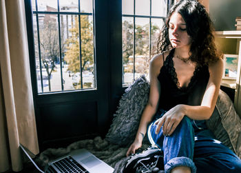 Young woman using mobile phone while sitting on window