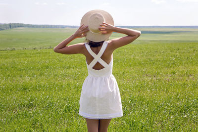 Young woman with long dark hair standing rear on green field. white dress and straw hat. 