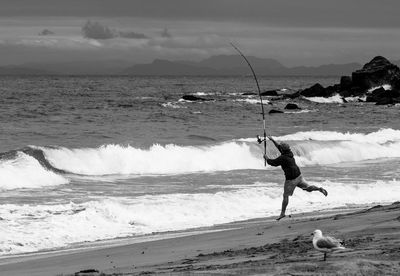 Full length of man holding fishing rod while jumping on beach against sky