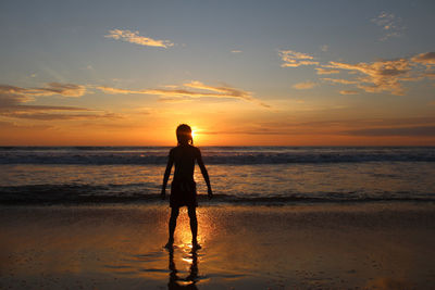 Boy standing at beach against sky during sunset