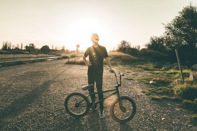 Young man with bicycle standing on field against clear sky during sunset