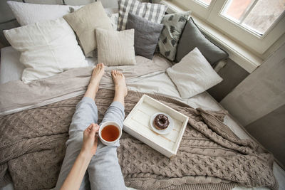 From above shot anonymous barefoot female enjoying tea donut while sitting comfortable bed morning