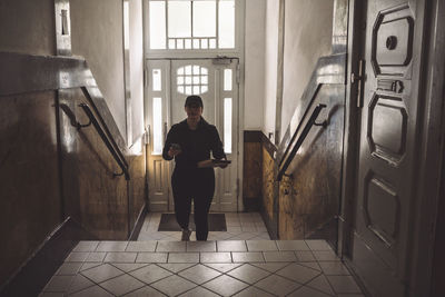 Portrait of delivery woman with phone and package climbing stairs against door