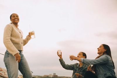 Happy woman giving drink to female friends against sky during picnic