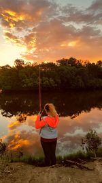 Rear view of woman doing fishing while standing by calm lake during sunset