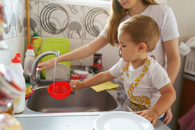 Close-up of baby boy with mother washing bowl in kitchen