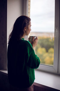 Woman in a green sweater holds a metal mug of cocoa and marshmallows