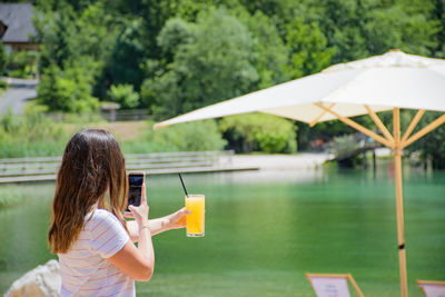 Woman photographing drink in glass with mobile phone by lake