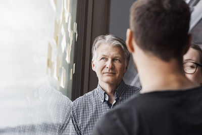 Businessman looking at adhesive notes while standing with colleagues by window