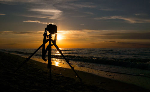 Silhouette camera on beach against sky during sunset