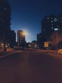 Empty road by illuminated buildings against sky at dusk