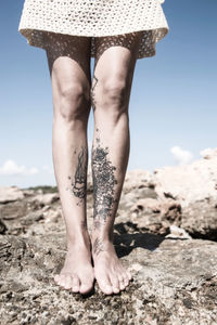 Low section of woman with tattooed legs standing on rock against sky