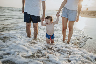 Midsection of mother and father with son standing in water at beach during sunset