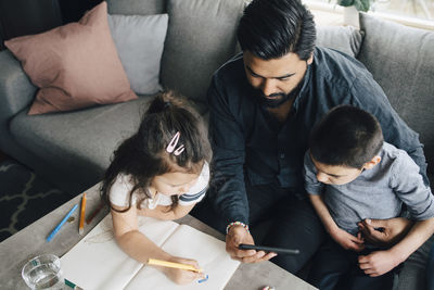 High angle view of father showing mobile phone to son while daughter drawing in book at home