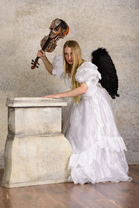 Young woman in angel costume holding violin against wall