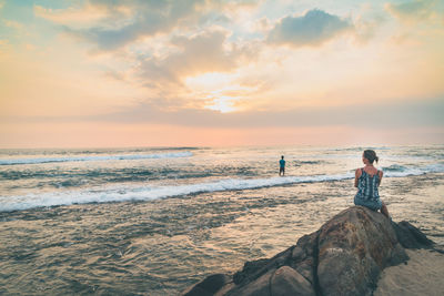 Woman sitting on rock at beach against sky during sunset