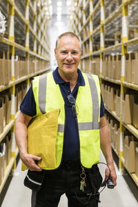 Portrait of smiling senior male worker holding package while standing on aisle at distribution warehouse
