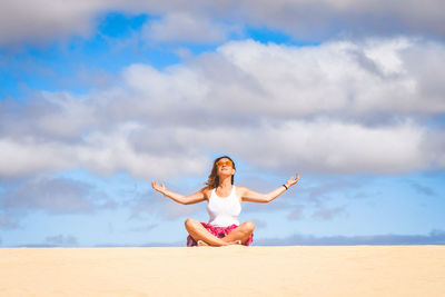 Full length of mature woman meditating while sitting on sand against cloudy sky during sunny day