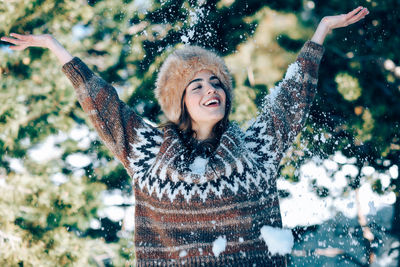 Cheerful young woman throwing snow while standing against trees during winter