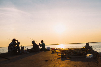 People fishing in sea while sitting on pier against sky during sunset