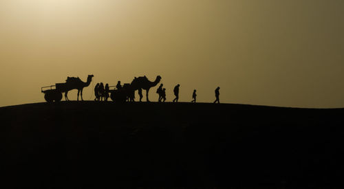 Silhouette people and camels on desert against sky during sunset