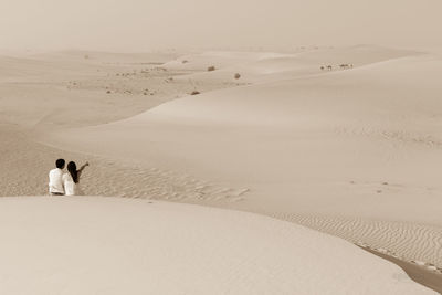 Rear view of couple sitting on sand at desert during sunny day