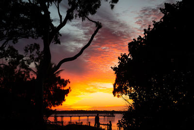 Silhouette trees by lake against orange sky