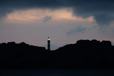 Silhouette lighthouse by building against sky during sunset