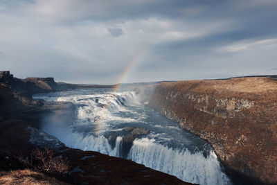 Scenic view of rainbow over waterfall against cloudy sky