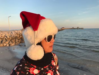 Mature woman wearing sunglasses and santa hat while standing at beach against sky during sunset