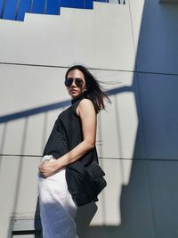 Portrait of young woman wearing sunglasses standing against wall