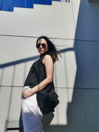 Portrait of young woman wearing sunglasses standing against wall