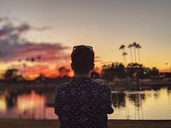 Rear view of man standing by lakeshore against sky during sunset