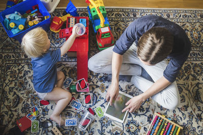 High angle view of blond baby boy playing toys by father using digital tablet in living room at home