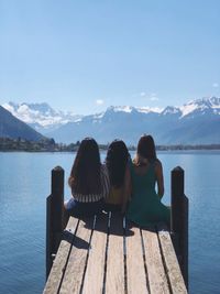 Rear view of friends sitting on pier over lake against blue sky